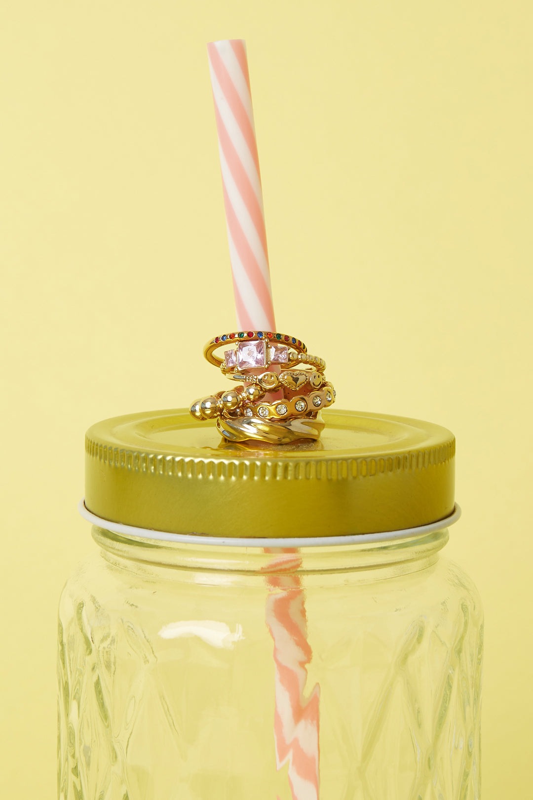 Stack of multiple gold-tone rings, including smiley face and heart designs, displayed on a pink and white striped straw inserted in a yellow-lidded glass jar with a yellow background