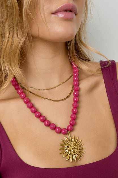Close-up of woman wearing vibrant berry pink beaded necklace with gold sun pendant against plum top