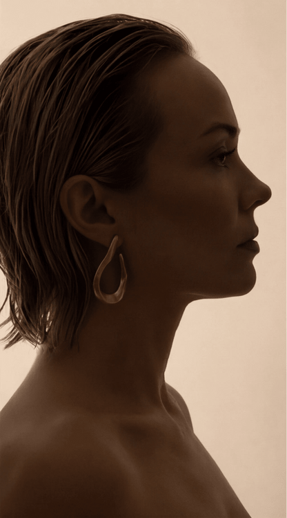 A woman wears a Glitch Core gold earring featuring an organic, teardrop-shaped open design on a silhouette portrait.