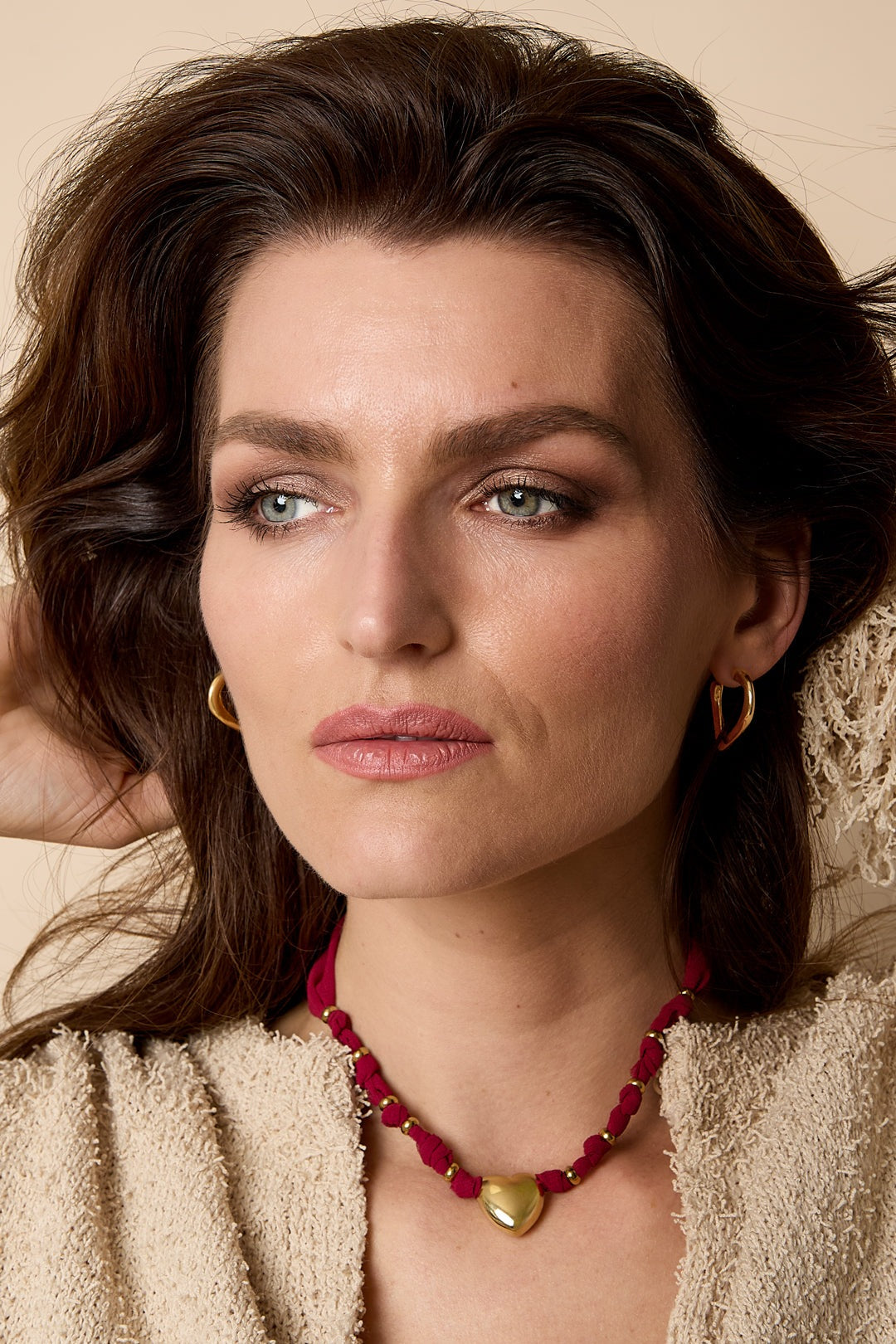 Woman wearing a gold heart pendant necklace with burgundy braided fabric against a beige background