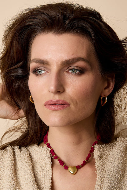 Woman wearing a gold heart pendant necklace with burgundy braided fabric against a beige background