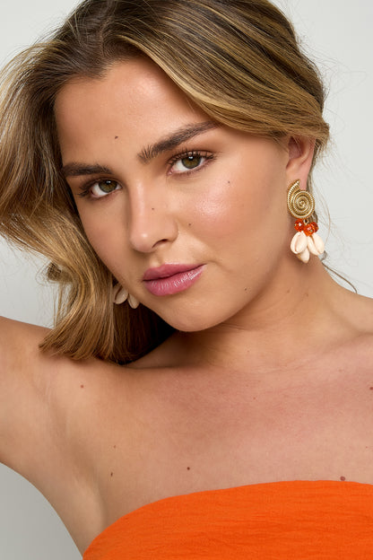 Close-up portrait of woman wearing gold spiral shell earrings with pink glass beads and white cowry shells, dressed in orange top