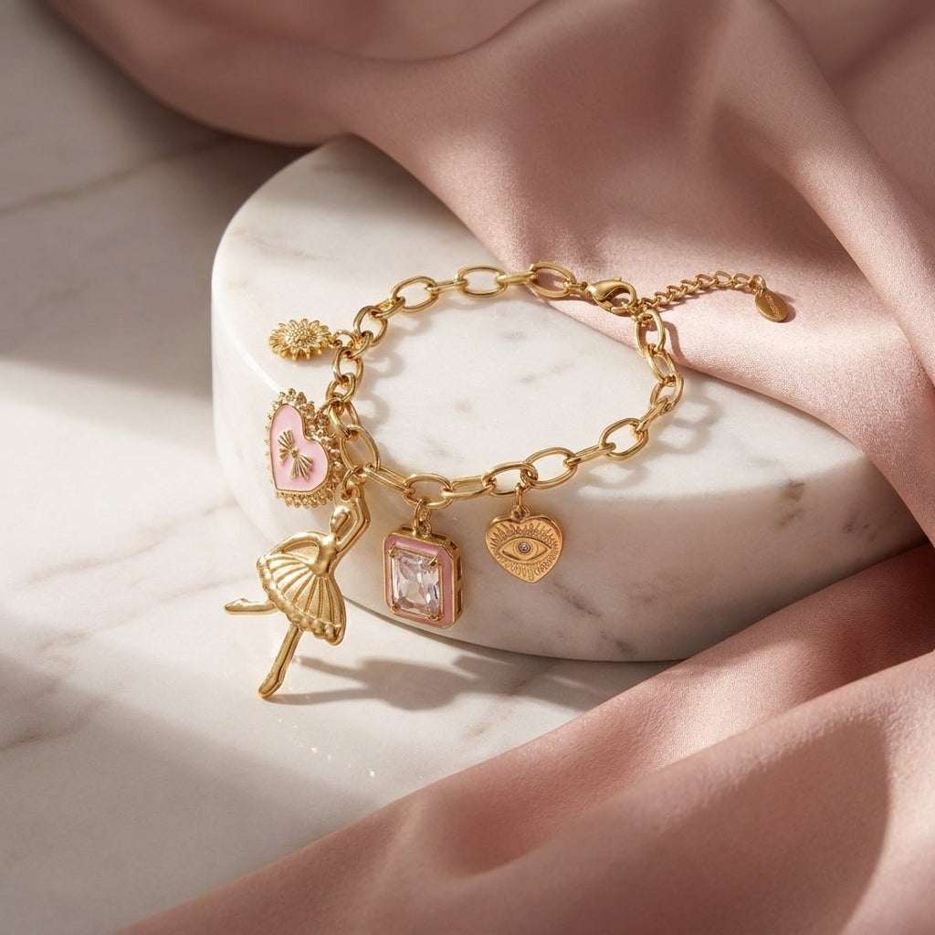 Gold chain bracelet with ballerina, heart, evil eye, and crystal charms on a marble stand with pink silk fabric.