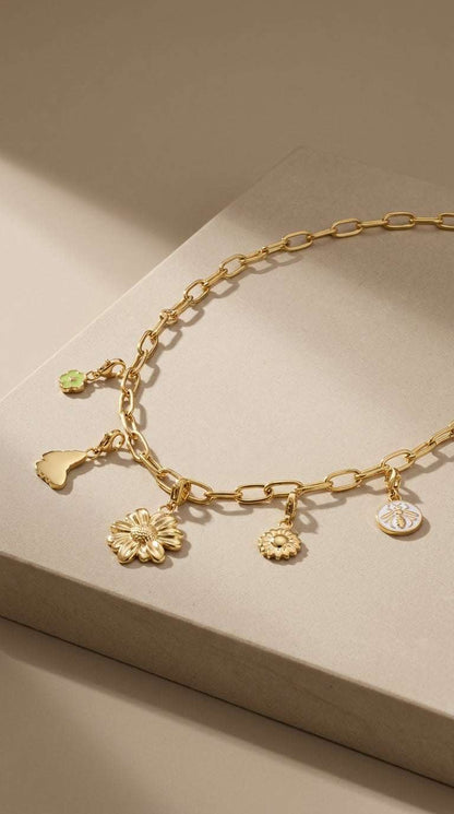Gold chain necklace featuring charms of flowers, a butterfly, and a bee, set on a neutral textured stone surface.