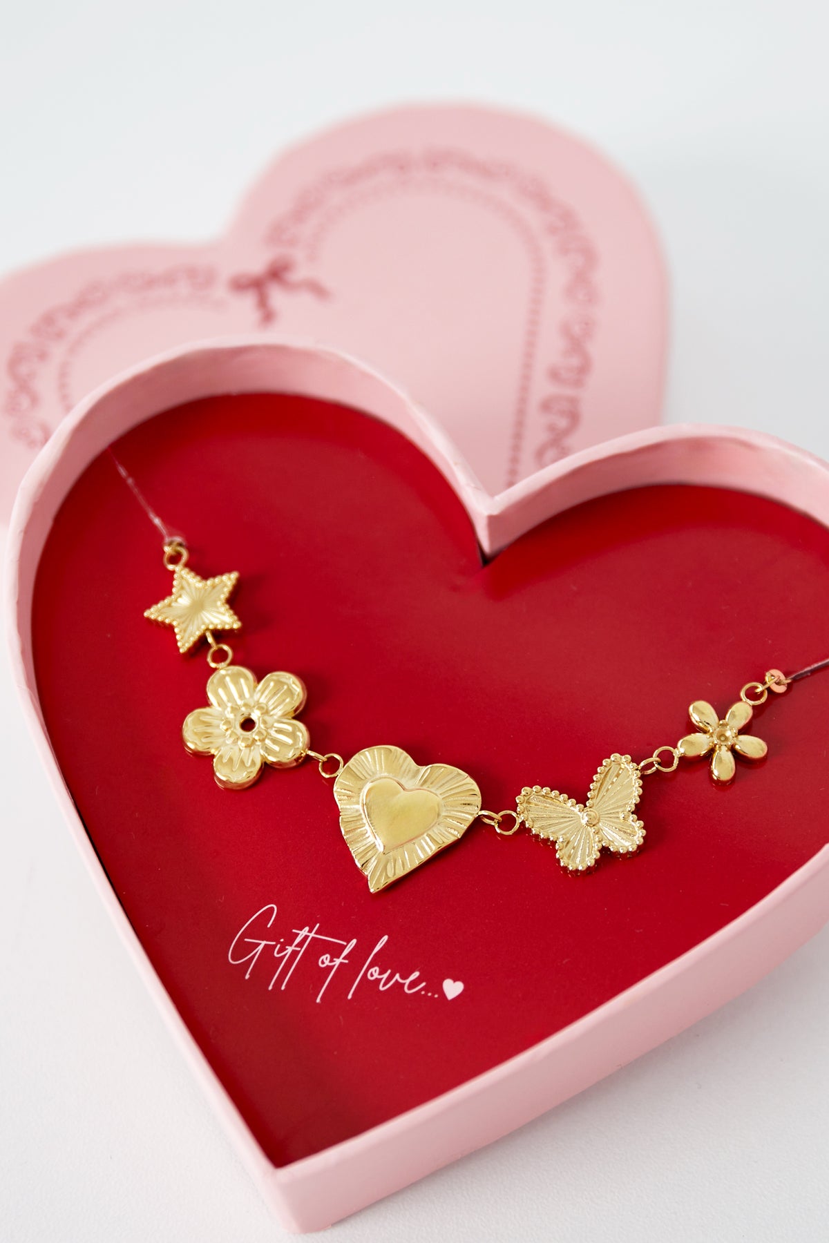 Close-up of gold pendant necklace featuring star, flower, heart, butterfly, and flower charms on gold chain on red card inside pink heart-shaped gift box with decorative lid