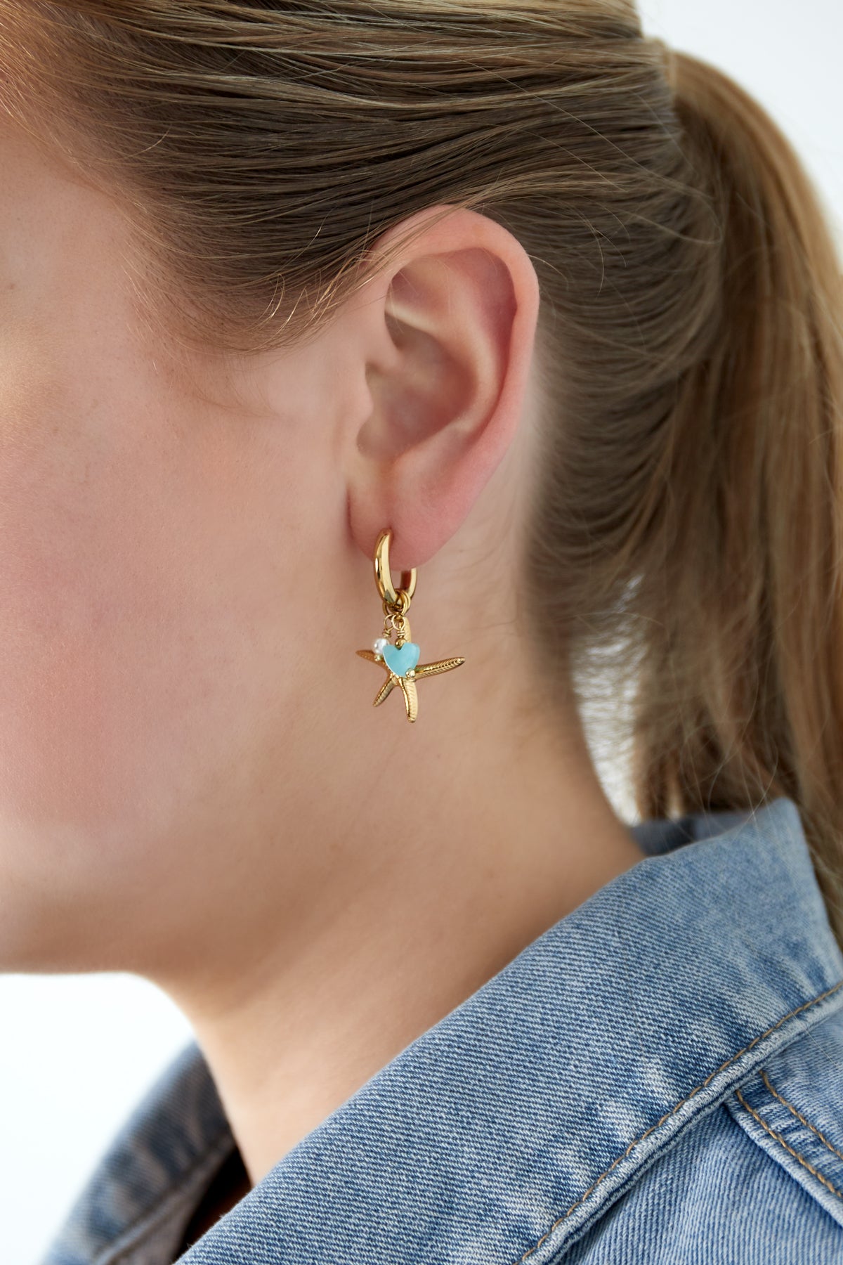 Close-up of woman wearing gold starfish charm hoop earring with aqua heart and pearl bead, styled with blue jean jacket
