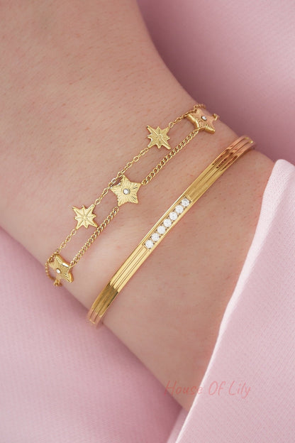 Close-up of wrist wearing gold star charm bracelet paired with a sleek gold cuff bracelet on pastel pink background