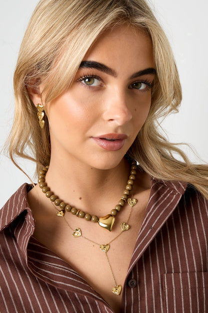 Side angle of model wearing olive bead necklace and layered necklaces with heart pendants, styled with a brown striped top