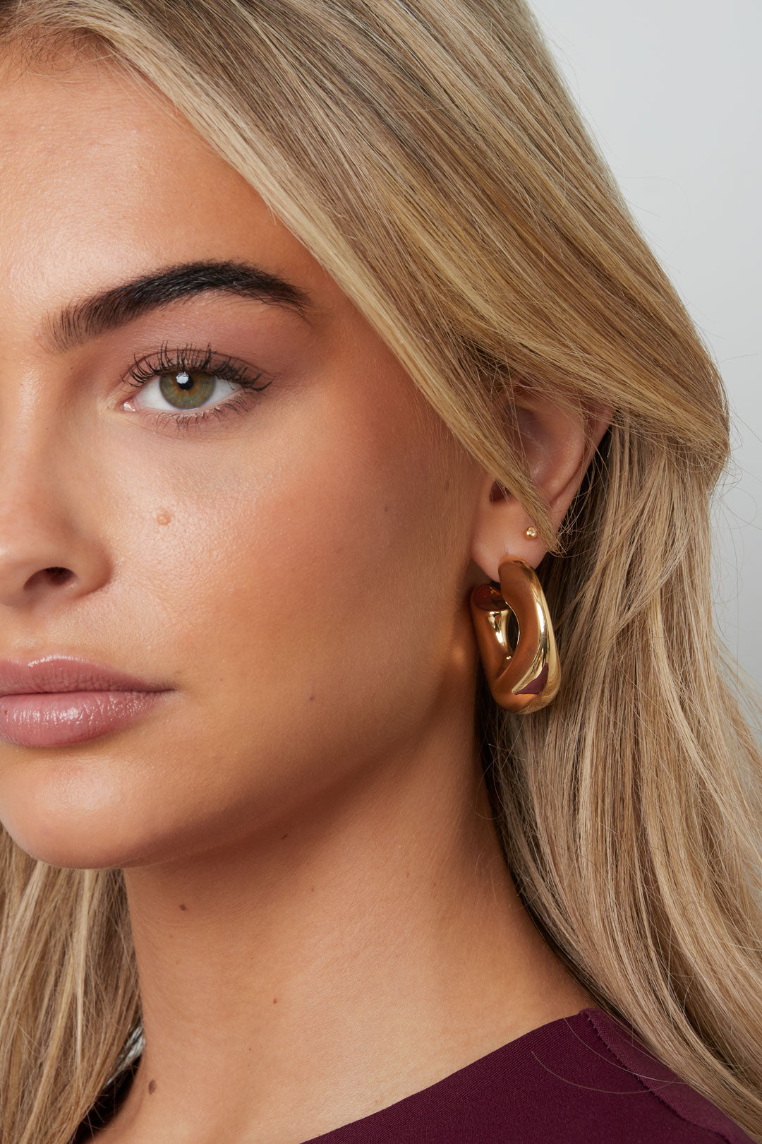 Close-up of a woman wearing chunky gold sculptural hoop earrings and a burgundy top