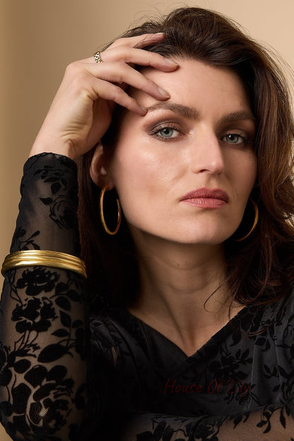 Woman wearing gold hoop earrings, a matching gold bangle, and a black floral sheer top