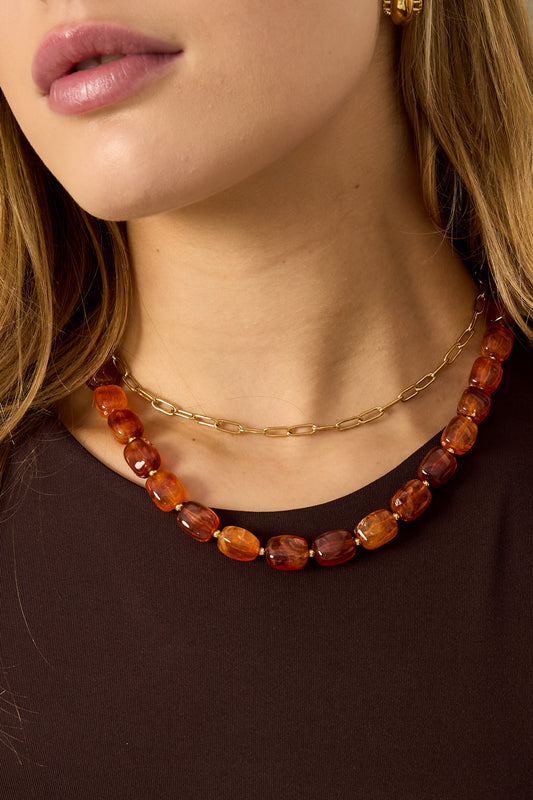 Close-up of woman wearing layered gold chain necklace and chunky red-orange acrylic beaded necklace over brown top