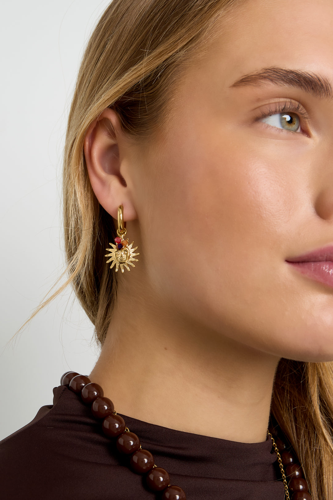 Close-up of a woman wearing gold sun charm hoop earring with amber beads styled with brown beaded necklace and dark brown top