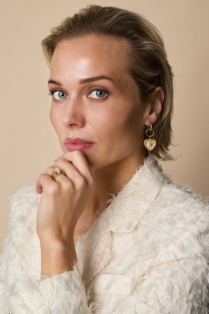 Model facing forward wearing gold heart charm drop earrings and white textured jacket with soft makeup