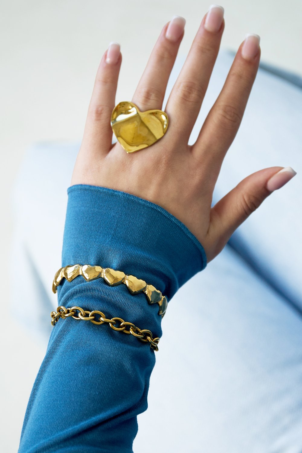 Heart Dent Era gold ring with a large, sculptural, indented heart shape, worn with two gold bracelets on blue sleeve.