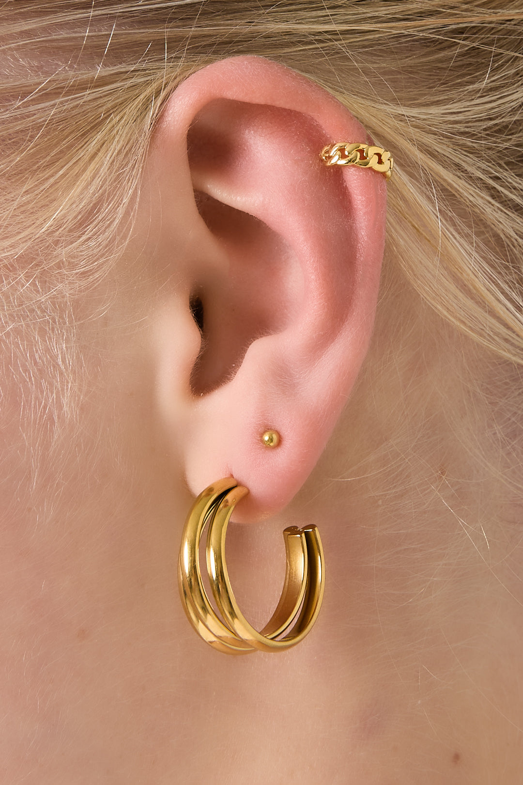 Model wearing a gold chunky chain ear cuff on the upper ear, paired with gold hoop earrings and a tiny gold stud.