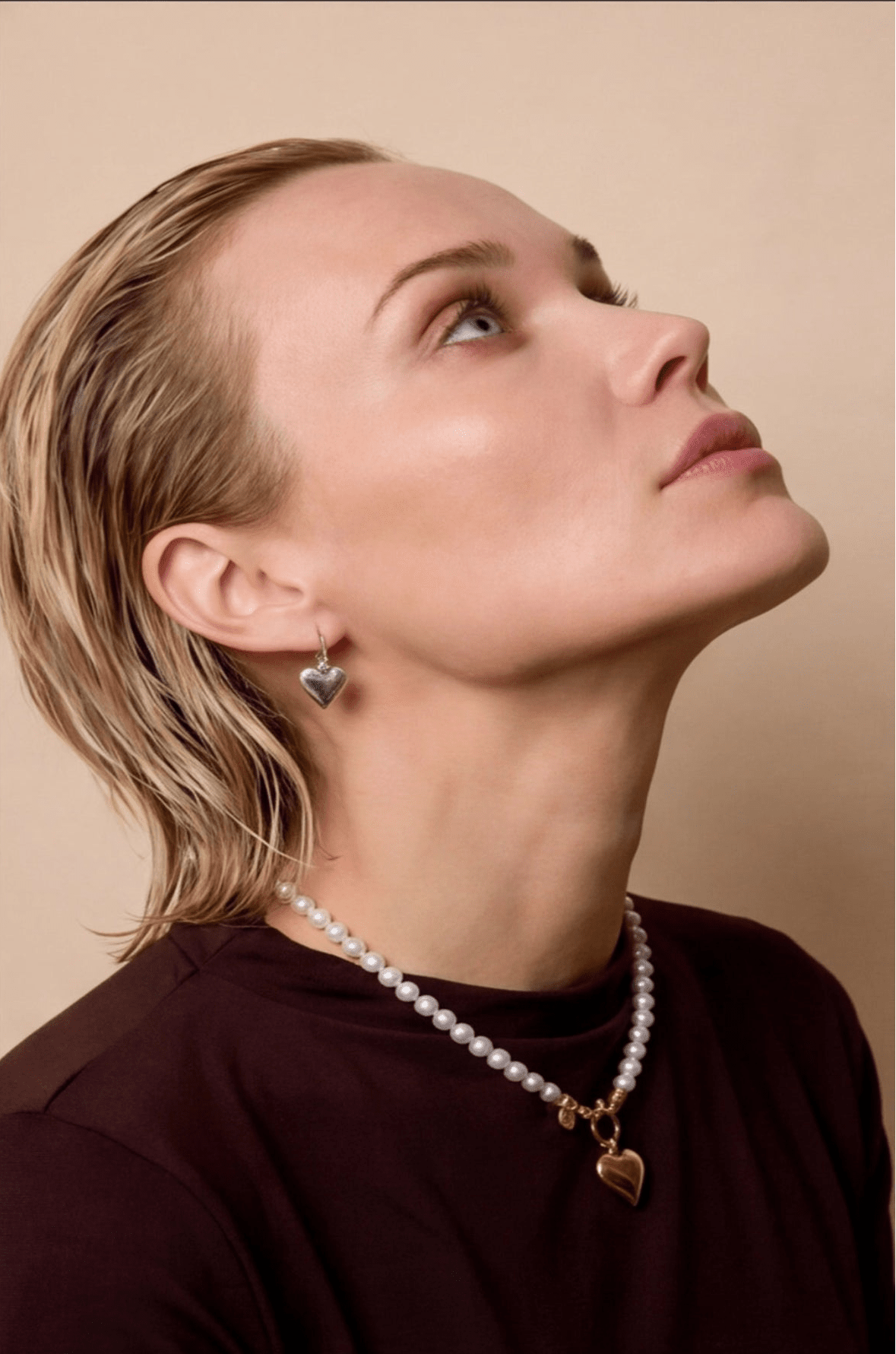 Woman wearing a pearl necklace and heart-shaped earrings against a beige background