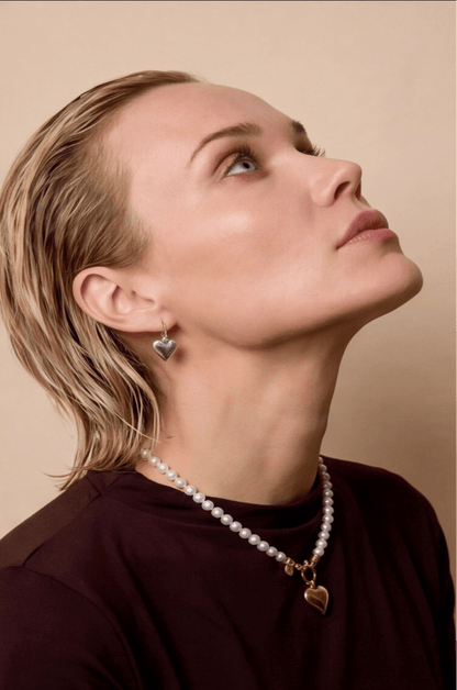 Woman wearing a pearl necklace and heart-shaped earrings against a beige background