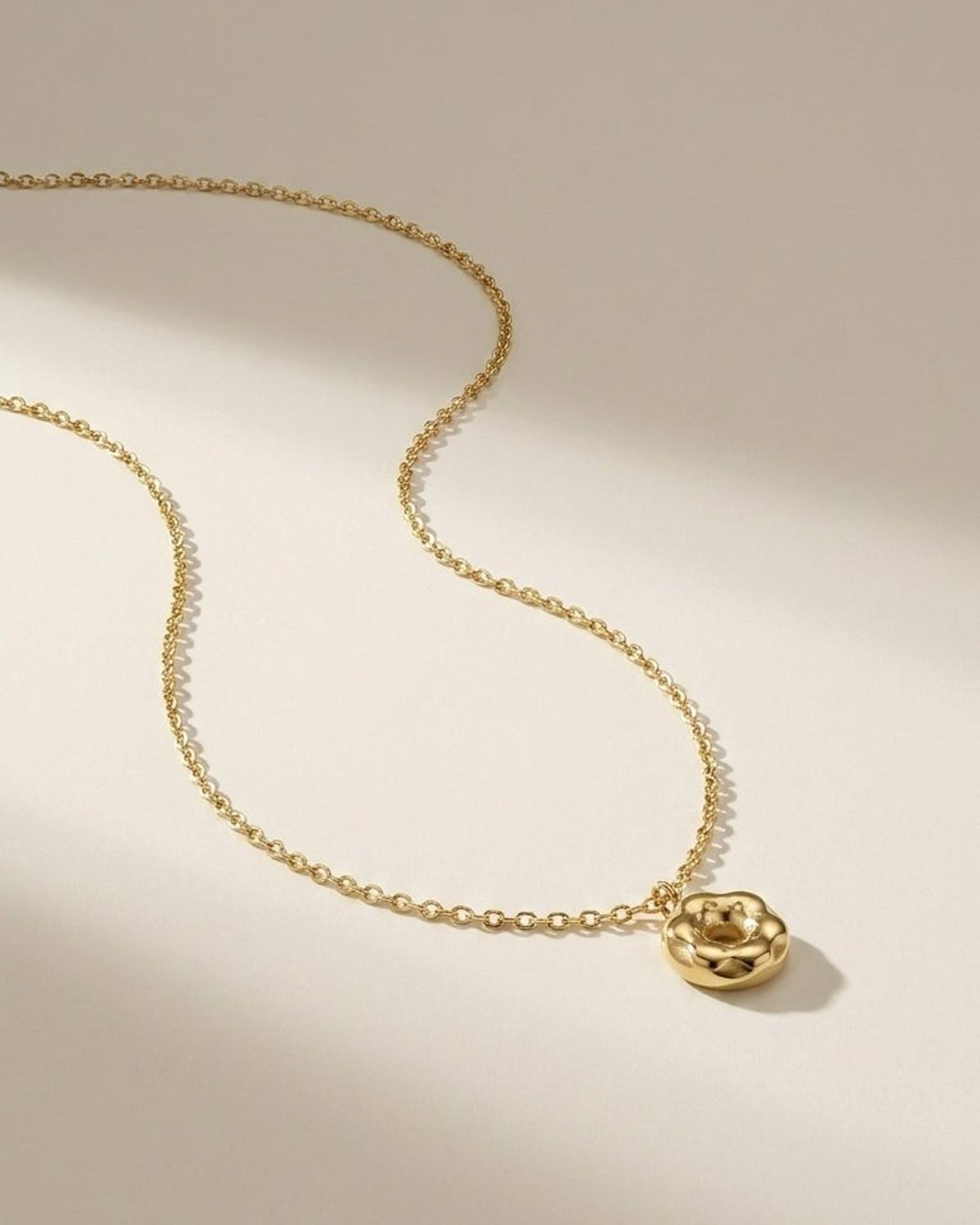 A gold chain necklace with a small, rounded donut-shaped pendant rests on a neutral surface.