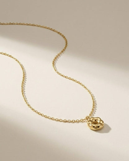 A gold chain necklace with a small, rounded donut-shaped pendant rests on a neutral surface.