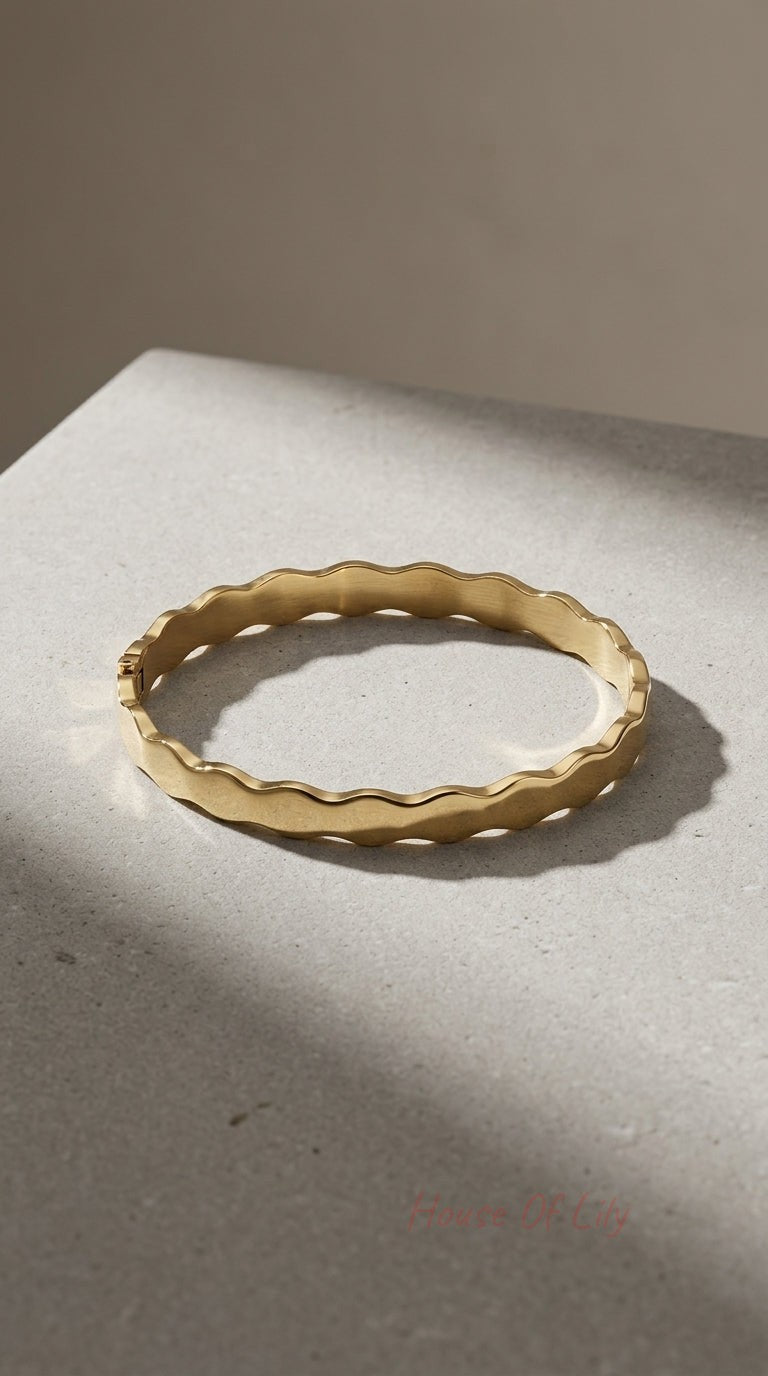 Gold hinged bangle with a wavy, scalloped edge and a brushed matte finish, resting on a textured grey surface.