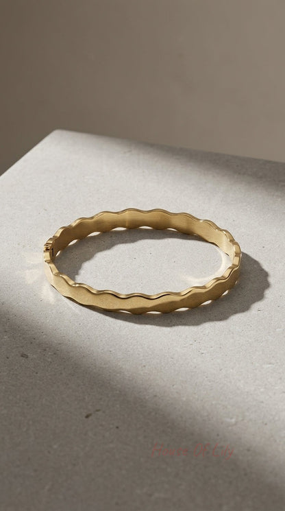 Gold hinged bangle with a wavy, scalloped edge and a brushed matte finish, resting on a textured grey surface.