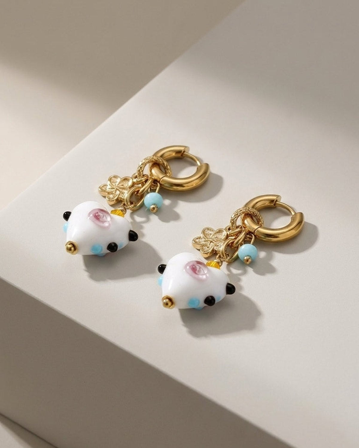 Gold hoop earrings with white heart-shaped charms, flower pendants, and small blue, black, and gold beads.