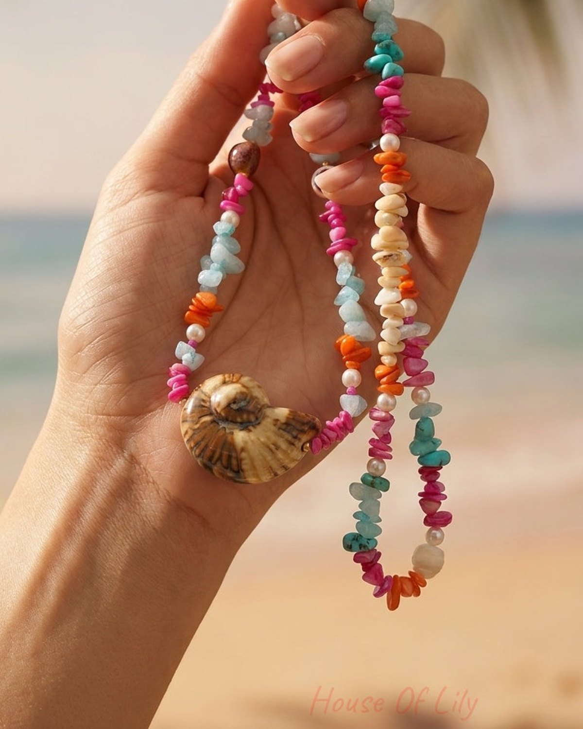 Hand holding a beaded shell necklace with colorful stone chips and pearls on a beach.