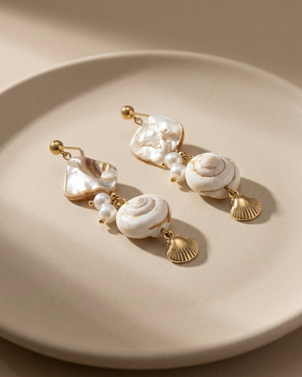 Gold-tone drop earrings featuring white shell accents, pearls, and small metal scallop charms on a beige plate.