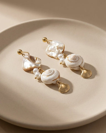 Gold-tone drop earrings featuring white shell accents, pearls, and small metal scallop charms on a beige plate.