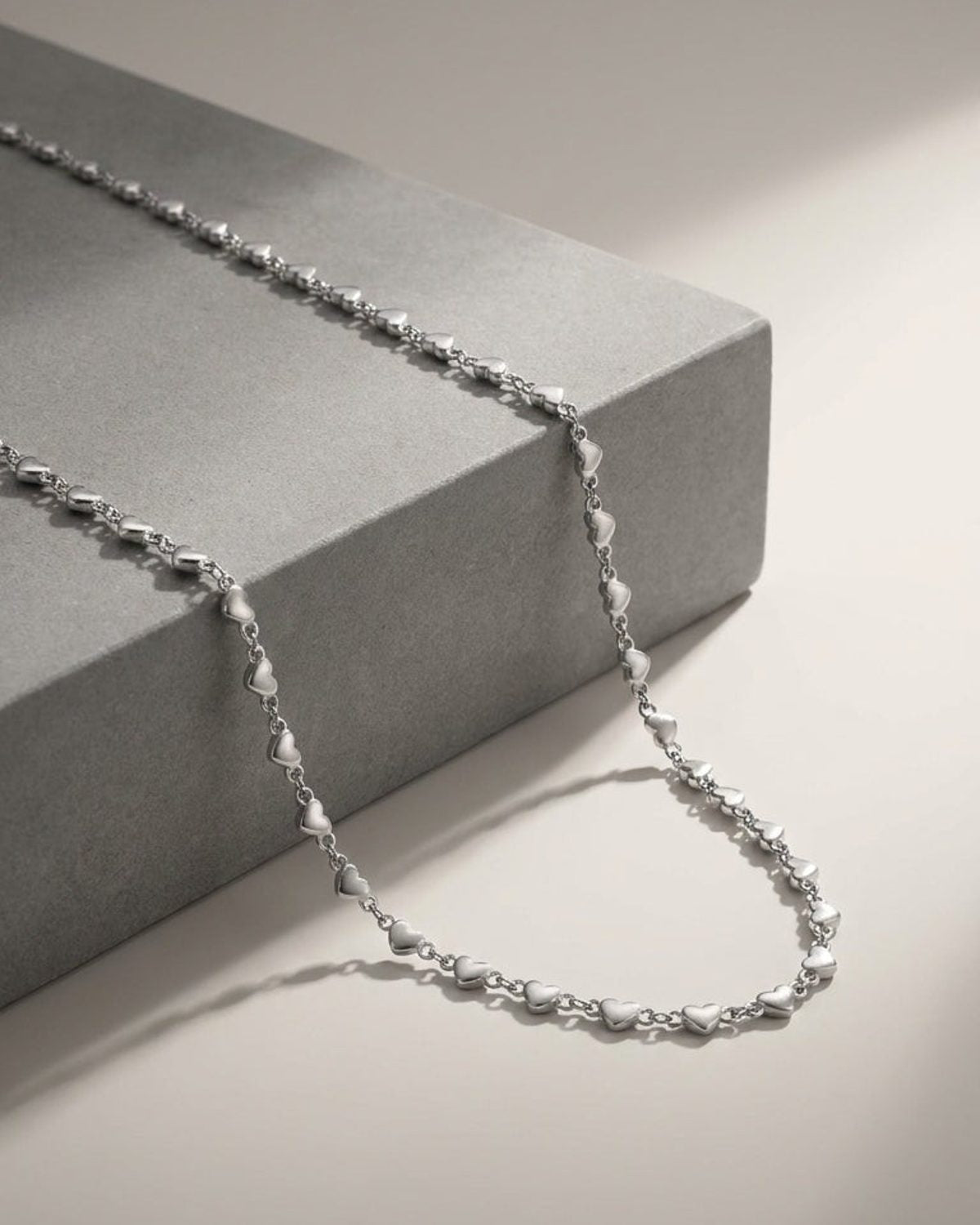 Delicate silver chain necklace featuring multiple small heart charms, draped over a grey block.