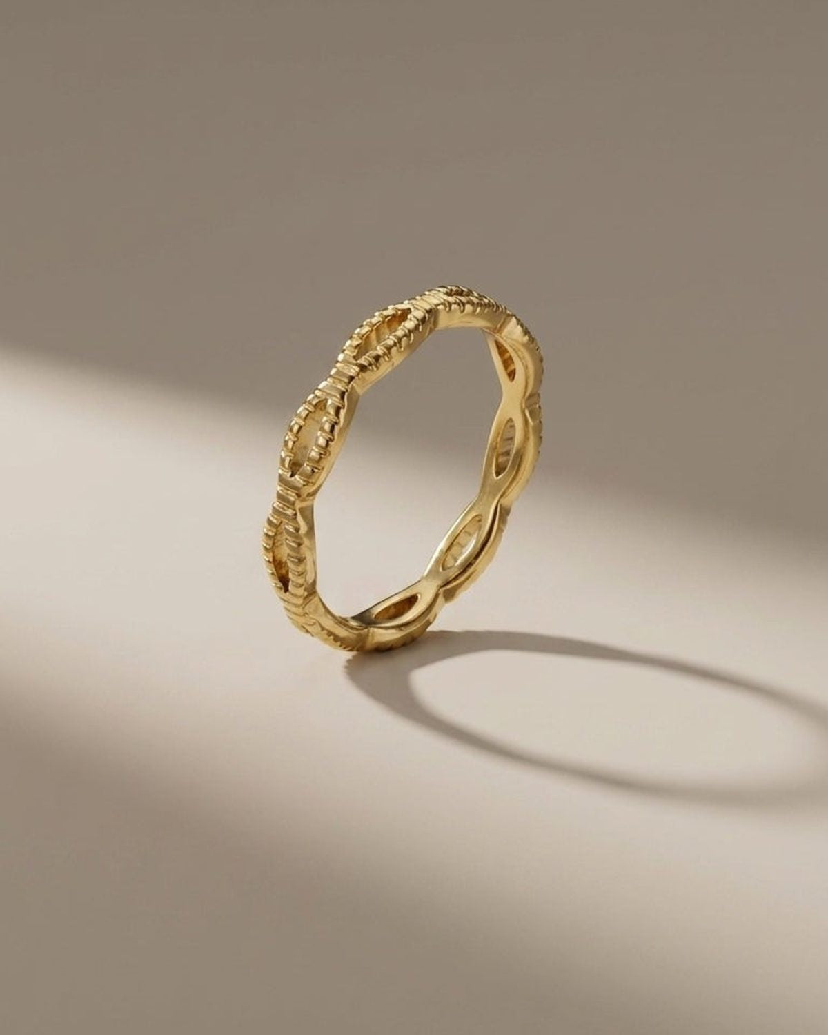 A thin gold ring with an openwork braided or looped design stands upright on a neutral surface.