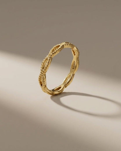 A thin gold ring with an openwork braided or looped design stands upright on a neutral surface.