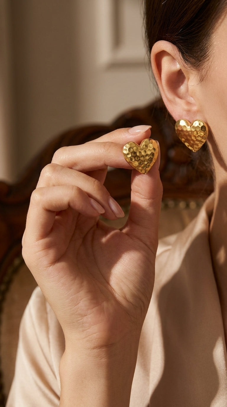 Gold-toned stud earrings in a heart shape with a textured, hammered finish being held by a hand.