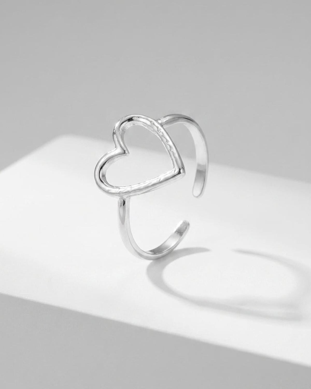 A silver open-band ring featuring an asymmetrical heart outline rests on a white surface.