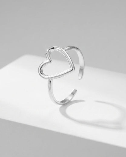 A silver open-band ring featuring an asymmetrical heart outline rests on a white surface.