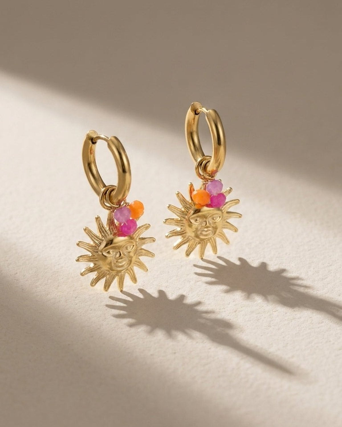Gold hoop earrings with dangling sun face charms, accented by small pink and orange bead clusters.