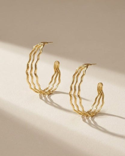 Two gold hoop earrings featuring a triple-band wavy design, displayed upright on a light surface.