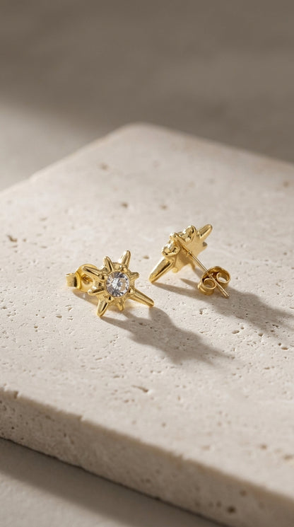 Gold-plated starburst stud earrings with a central clear cubic zirconia stone on a textured stone surface.