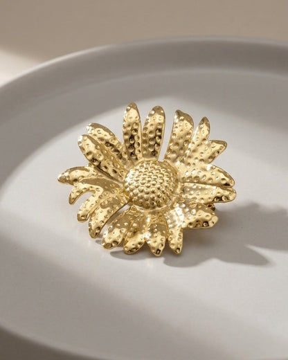Gold sunflower brooch with a textured, hammered finish rests on a grey dish.