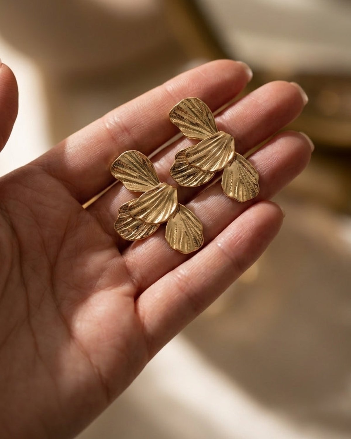 Gold flower drop earrings featuring textured petal shapes, held in a person's open palm.