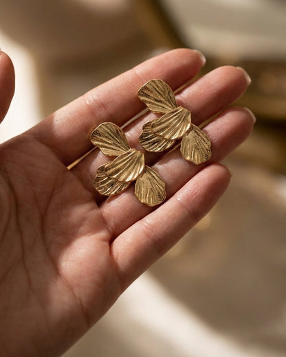 Gold flower drop earrings featuring textured petal shapes, held in a person's open palm.