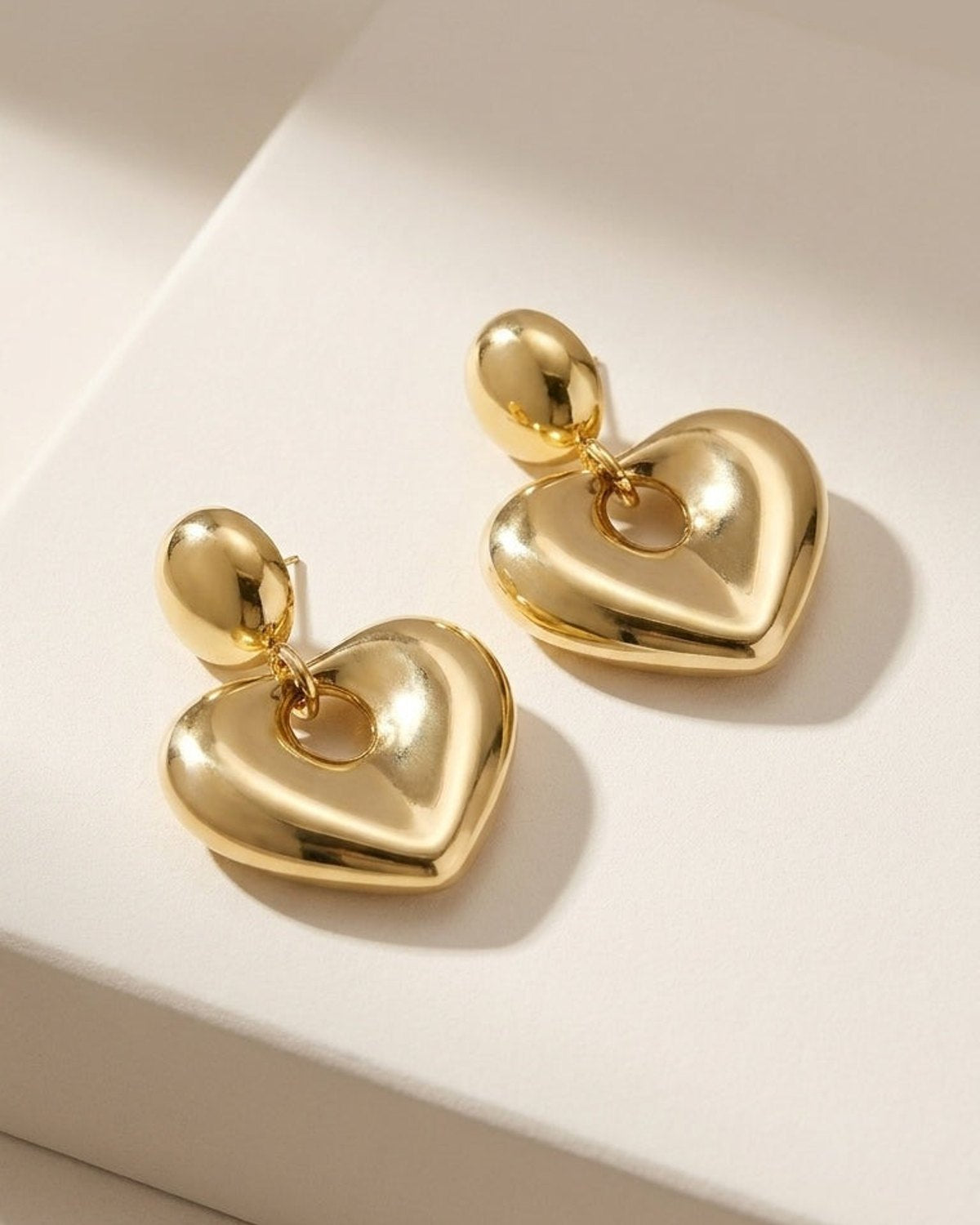Polished gold drop earrings featuring a domed oval stud attached to a puffy, hollow heart pendant.
