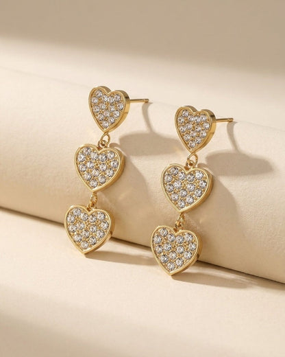 Gold-tone triple heart dangle earrings encrusted with pavé clear rhinestones.