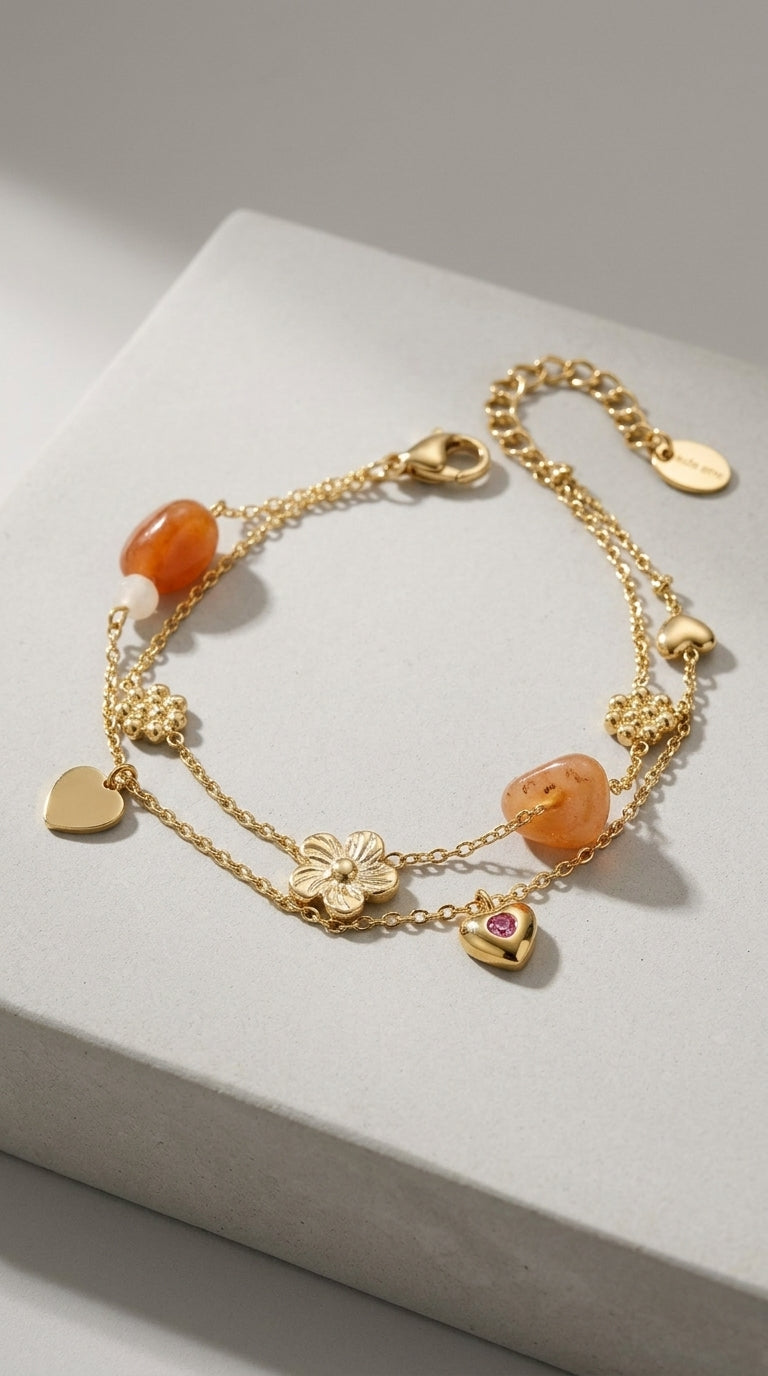 Gold chain charm bracelet with orange stones, flower charms, and a heart with a pink gem on a grey surface.