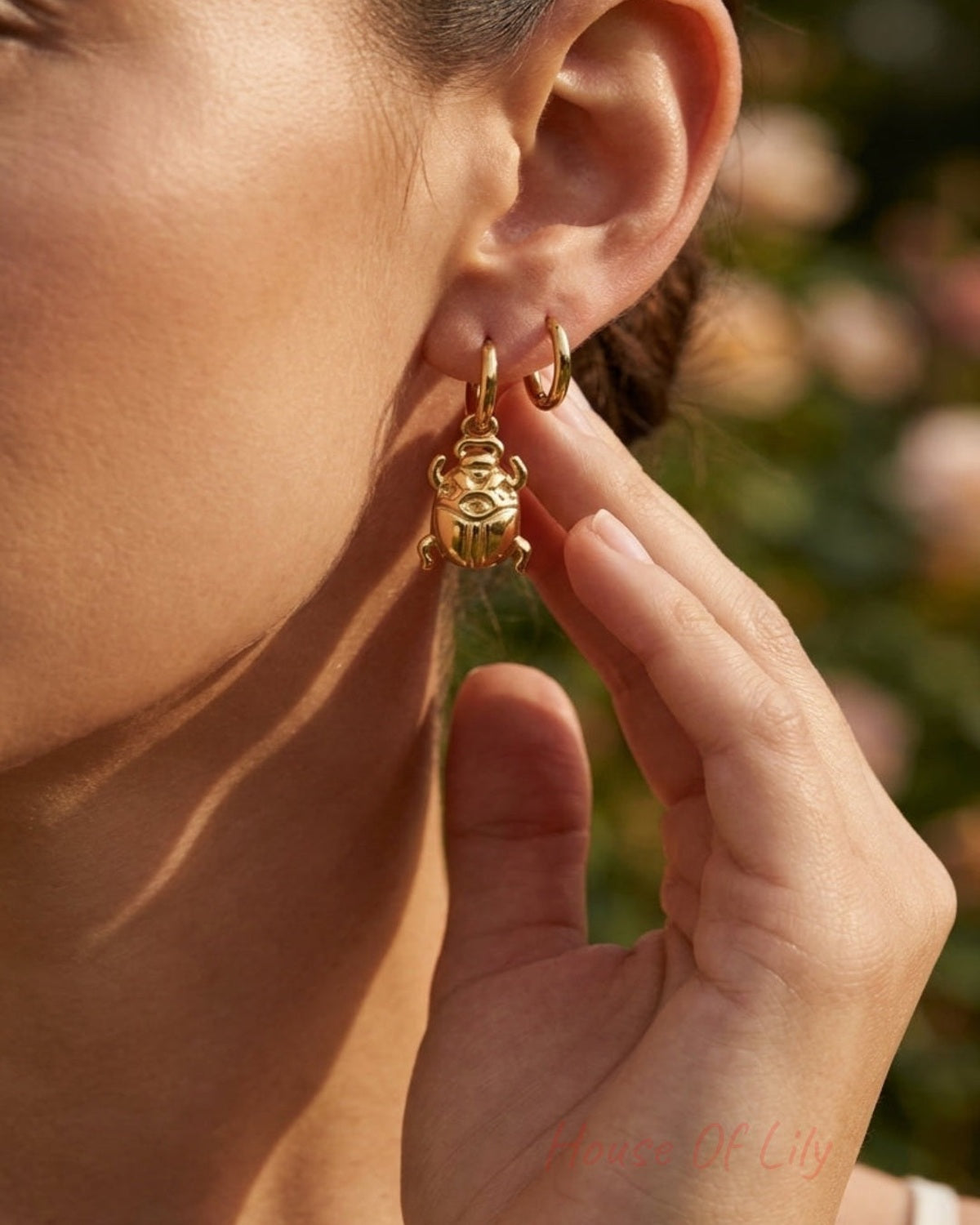 A close-up lifestyle shot of a woman wearing a small gold huggie hoop earring with a dangling gold monkey charm, set against a soft-focus outdoor background.