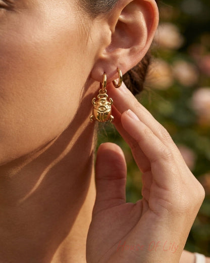 A close-up lifestyle shot of a woman wearing a small gold huggie hoop earring with a dangling gold monkey charm, set against a soft-focus outdoor background.