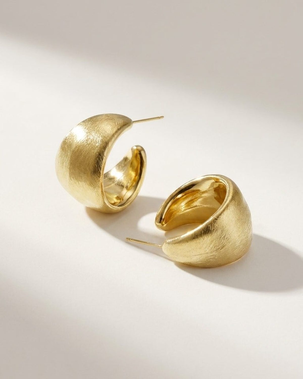 Chunky gold hoop earrings with a brushed metal finish and classic post backs, resting on a neutral surface.