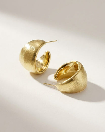 Chunky gold hoop earrings with a brushed metal finish and classic post backs, resting on a neutral surface.