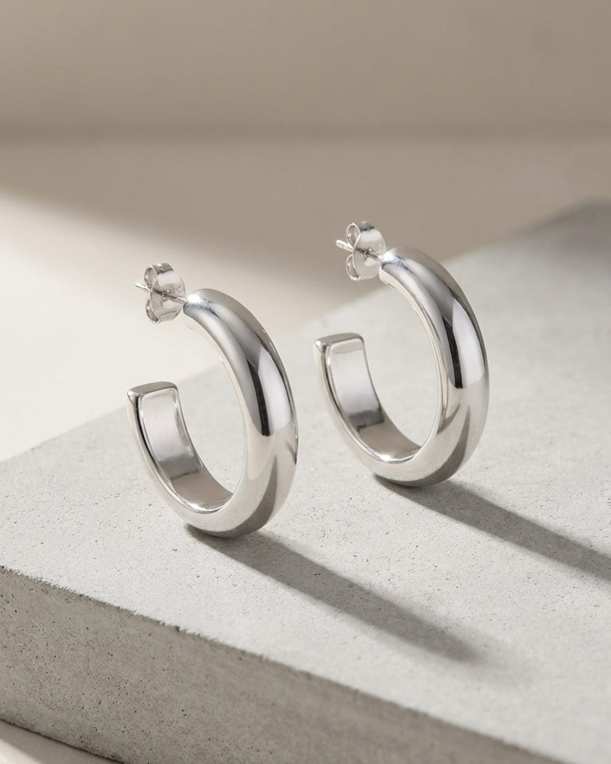 Minimalist silver hoop earrings with a thick, polished, chunky design on a grey stone surface.