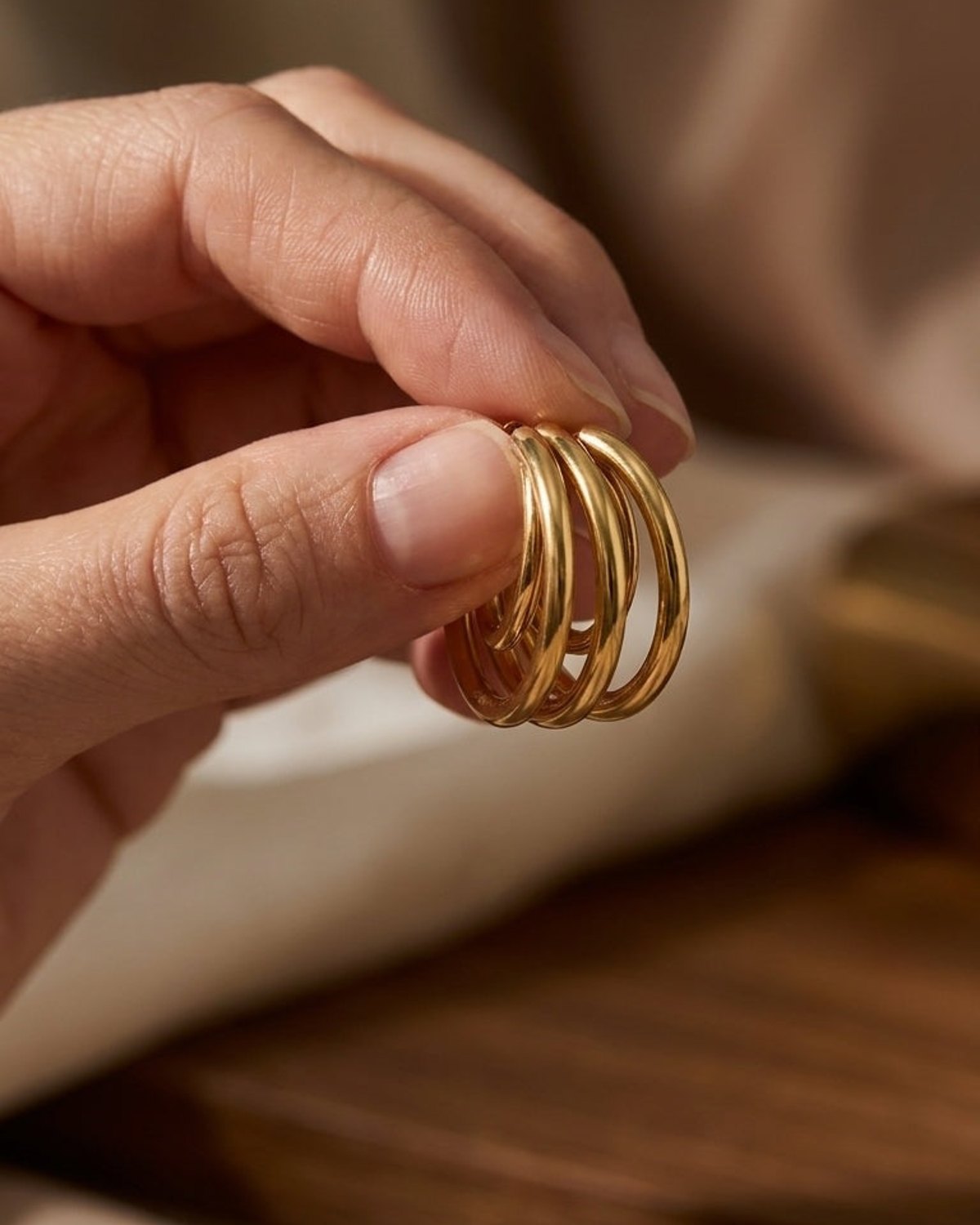 A hand holds up a cluster of four polished gold hoop earrings against a neutral background.